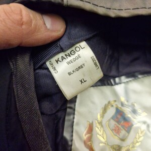 Może przedstawiać: Zbliżenie czarnej i szarej czapki Kangol Wedge. Etykieta głosi "KANGOL WEDGE BLK/GREY XL". Czapka ma ciemnoszarą powierzchnię zewnętrzną i granatowe wnętrze. Złoty emblemat jest widoczny na podszewce.