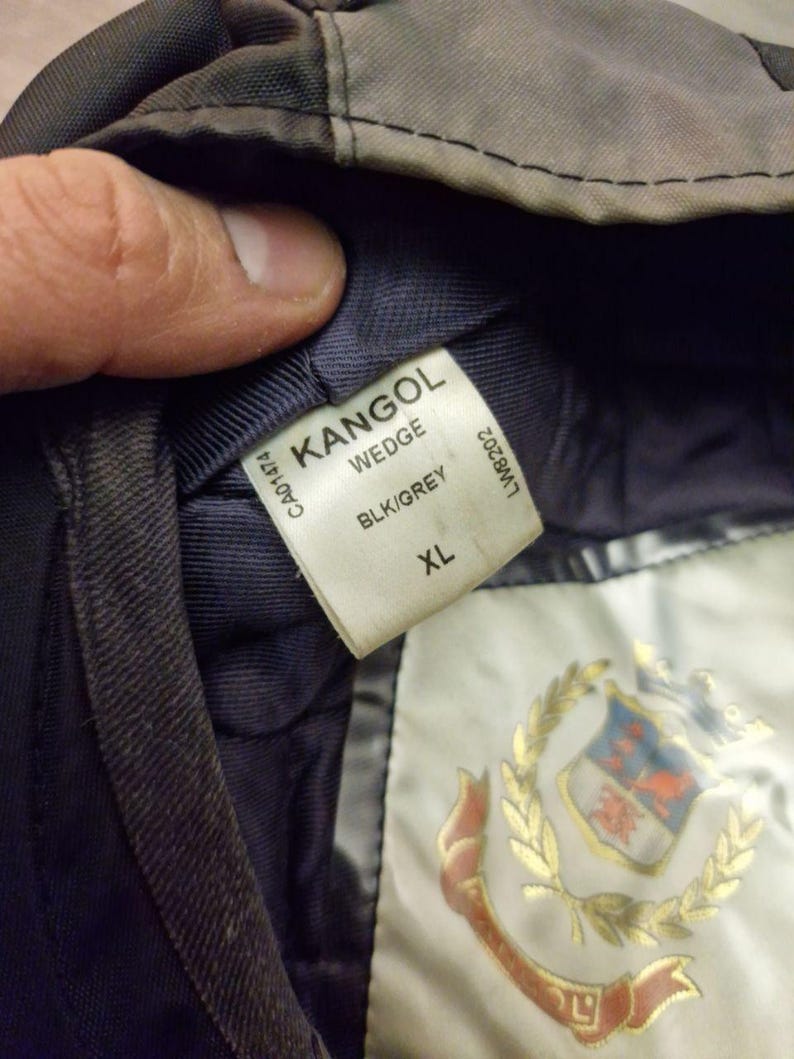 Może przedstawiać: Zbliżenie czarnej i szarej czapki Kangol Wedge. Etykieta głosi "KANGOL WEDGE BLK/GREY XL". Czapka ma ciemnoszarą powierzchnię zewnętrzną i granatowe wnętrze. Złoty emblemat jest widoczny na podszewce.