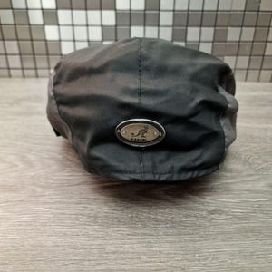 Może przedstawiać: Czarna i szara czapka Kangol z małą metalową plakietką z logo kangura. Czapka wykonana jest z wytrzymałej tkaniny i ma klasyczny, zaokrąglony kształt. Kapelusz został zaprojektowany z myślą o wygodnym dopasowaniu.