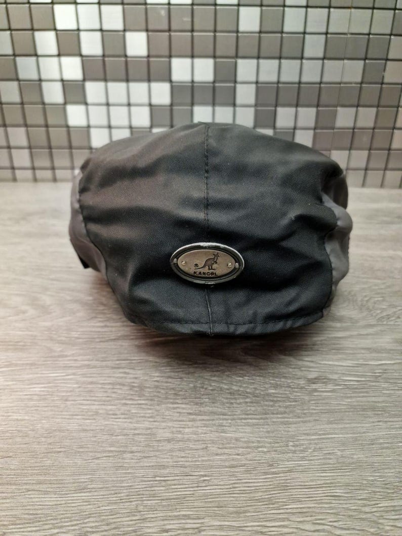 Może przedstawiać: Czarna i szara czapka Kangol z małą metalową plakietką z logo kangura. Czapka wykonana jest z wytrzymałej tkaniny i ma klasyczny, zaokrąglony kształt. Kapelusz został zaprojektowany z myślą o wygodnym dopasowaniu.