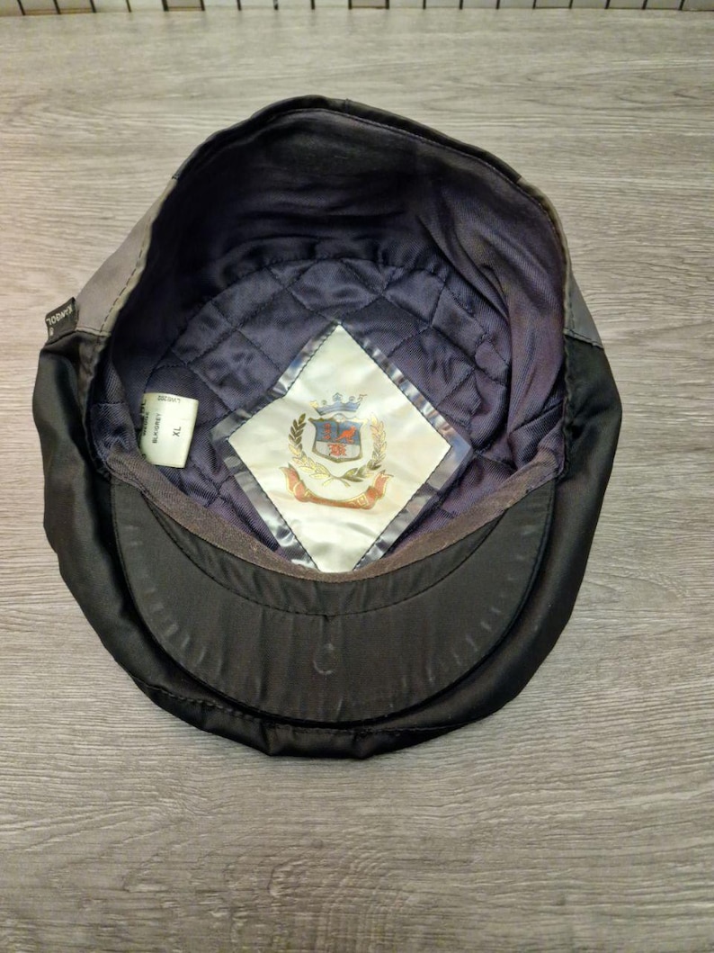 Może przedstawiać: Czarna i szara czapka Kangol, widziana od wewnątrz. Wnętrze posiada pikowaną granatową podszewkę i białą metkę z herbem. Czapka ma zakrzywiony daszek i strukturalny kształt.