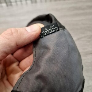 Może przedstawiać: Zbliżenie szarego i czarnego przedmiotu z tkaniny z małą czarną metką. Na metce znajduje się napis "showerproof" w białym skrypcie. Przedmiot wydaje się być wykonany z trwałego, wodoodpornego materiału.