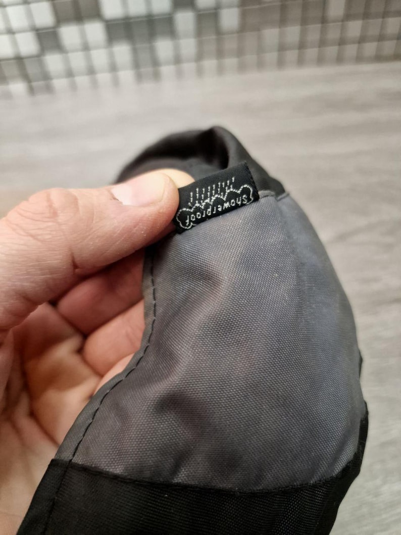 Może przedstawiać: Zbliżenie szarego i czarnego przedmiotu z tkaniny z małą czarną metką. Na metce znajduje się napis "showerproof" w białym skrypcie. Przedmiot wydaje się być wykonany z trwałego, wodoodpornego materiału.