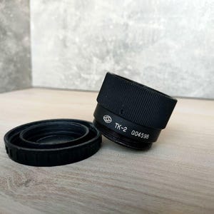 Puede incluir: Un objetivo de cámara negro con una tapa de objetivo a juego. El objetivo tiene el texto "TK-2 004598" impreso. La tapa del objetivo es redonda y tiene un borde estriado. El objetivo y la tapa están sobre una superficie de madera clara.