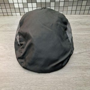 Może przedstawiać: Czarna czapka typu newsboy z szarymi panelami bocznymi. Czapka wykonana jest z gładkiego, prawdopodobnie syntetycznego materiału. Czapka leży na drewnianej powierzchni.