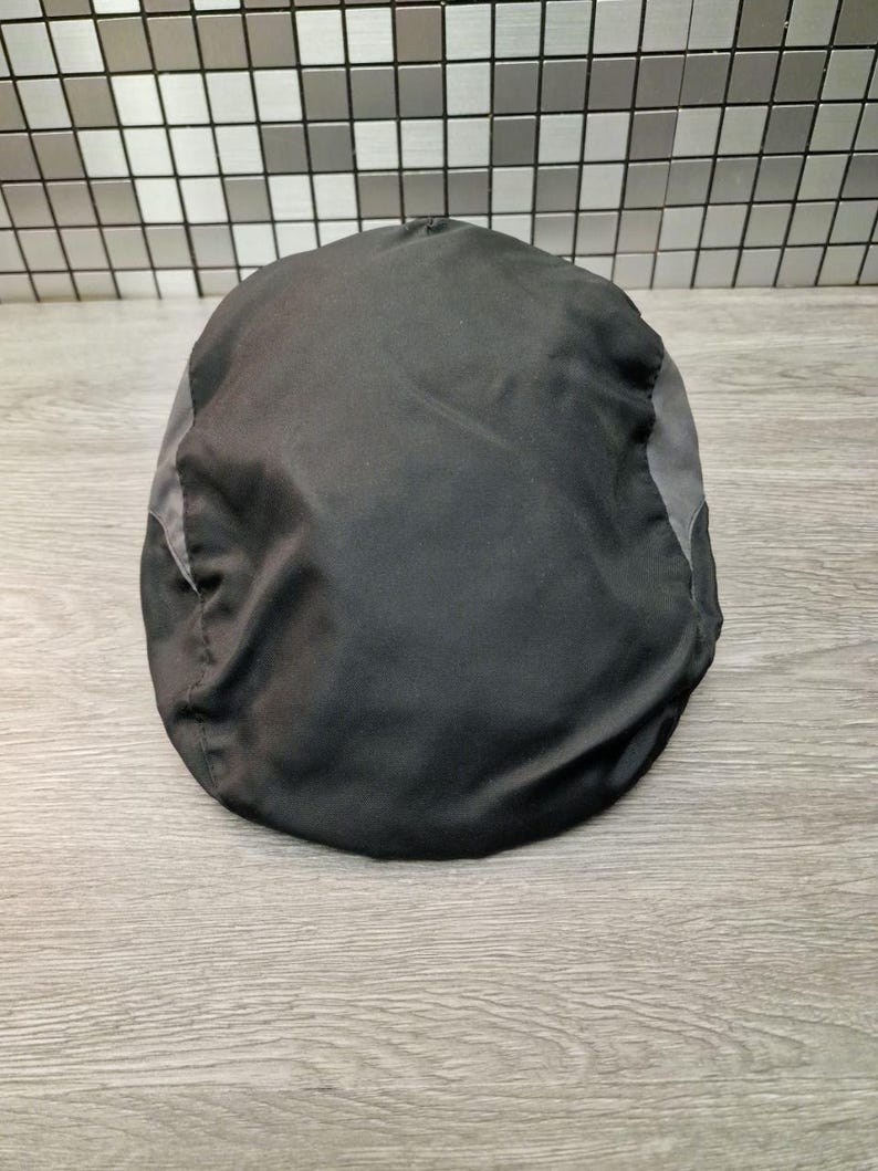 Może przedstawiać: Czarna czapka typu newsboy z szarymi panelami bocznymi. Czapka wykonana jest z gładkiego, prawdopodobnie syntetycznego materiału. Czapka leży na drewnianej powierzchni.