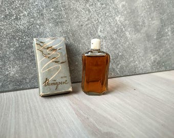 May include: Vintage perfume bottle with a white cap and a rectangular shape, filled with amber liquid. A matching box with the Cyrillic text "Bemepeк" and a gold leaf design is next to the bottle. The background is a gray wall and a wooden surface.