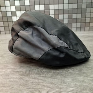 Może przedstawiać: Czarna i szara czapka typu newsboy. Czapka wykonana jest z gładkiego, prawdopodobnie wodoodpornego materiału. Projekt posiada ciemniejszy szary panel z przodu, przyszyty do czarnego korpusu. Czapka spoczywa na drewnianej powierzchni.