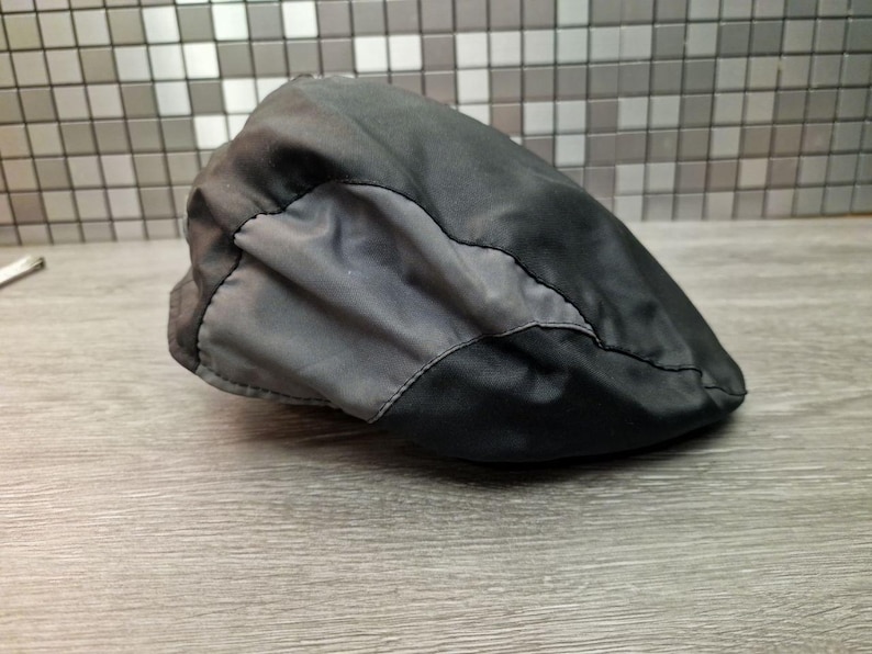 Może przedstawiać: Czarna i szara czapka typu newsboy. Czapka wykonana jest z gładkiego, prawdopodobnie wodoodpornego materiału. Projekt posiada ciemniejszy szary panel z przodu, przyszyty do czarnego korpusu. Czapka spoczywa na drewnianej powierzchni.