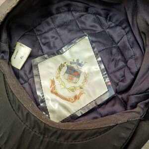 Może przedstawiać: Czarna czapka z pikowaną granatową podszewką. Wnętrze posiada etykietę z motywem herbu, złotymi liśćmi laurowymi i czerwonym bannerem. Czapka jest oznaczona rozmiarem XL.