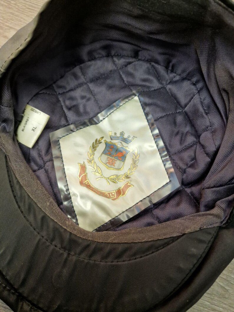 Może przedstawiać: Czarna czapka z pikowaną granatową podszewką. Wnętrze posiada etykietę z motywem herbu, złotymi liśćmi laurowymi i czerwonym bannerem. Czapka jest oznaczona rozmiarem XL.