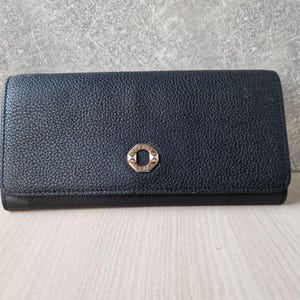 May include: A dark blue leather wallet with a textured surface. The wallet has a gold-colored, geometric clasp in the center. The wallet is rectangular and appears to be closed.
