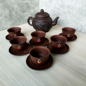 Op de afbeelding: Een bruine keramische theeset met een theepot met een drakenontwerp en zes bijpassende kopjes en schotels. De kopjes en schotels hebben een klein embleem. De theeset is gemaakt van donkerbruin keramisch materiaal.