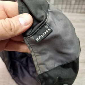 Może przedstawiać: Zbliżenie na czapkę Kangol, gł&oacute;wnie szarą i czarną. Czapka posiada małą, prostokątną metkę z białym logo Kangol. Czapka wydaje się być wykonana z wytrzymałego, prawdopodobnie wodoodpornego materiału.
