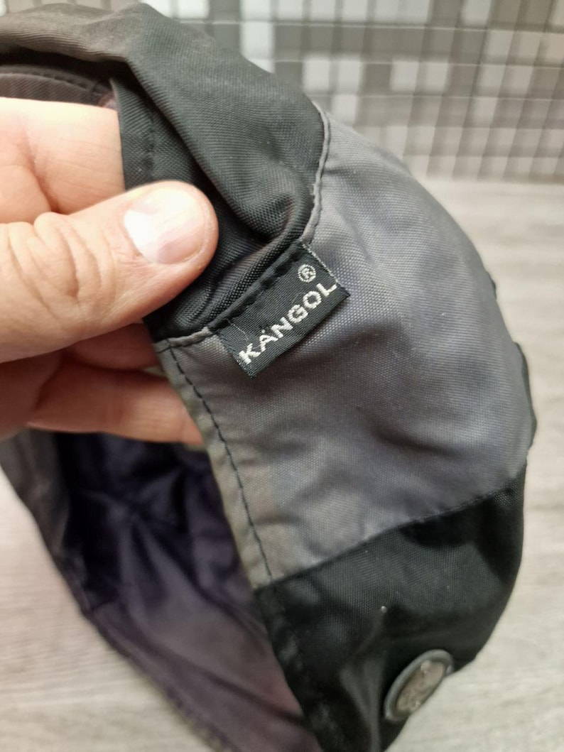 Może przedstawiać: Zbliżenie na czapkę Kangol, gł&oacute;wnie szarą i czarną. Czapka posiada małą, prostokątną metkę z białym logo Kangol. Czapka wydaje się być wykonana z wytrzymałego, prawdopodobnie wodoodpornego materiału.