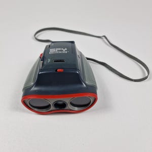 May include: A pair of black and gray Spy Gear binoculars with red accents around the lenses. The binoculars have a black strap and the text "SPY GEAR" is visible on the top.