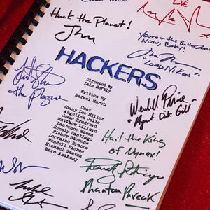 Op de afbeelding: Een spiraalgebonden script getiteld "HACKERS" met talrijke handtekeningen in verschillende kleuren. De titel is blauw, de handtekeningen in zwart, blauw en rood. De achtergrond is rood en glinsterend.