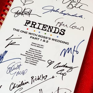 May include: A white script with the title "FRIENDS" and the text "THE ONE WITH ROSS'S WEDDING PART 1 & 2" is covered in signatures. The script is bound with a red spiral binding and rests on a red surface.