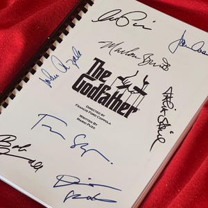 May include: A white page with the title "The Godfather" and the names of the director and writer. The page is signed by several people, including Al Pacino, Marlon Brando, and Robert Duvall.