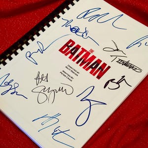 May include: A white script with the title "The Batman" in red and black lettering. The script is signed by multiple people in blue ink. The script is on a red background.