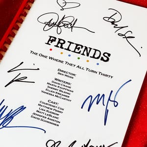 Puede incluir: Un guion blanco de Friends, titulado "The One Where They All Turn Thirty", con múltiples firmas en tinta negra y azul. El guion está encuadernado con una espiral roja y descansa sobre una superficie roja.