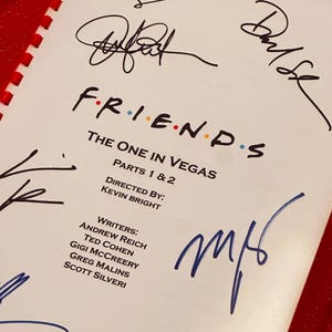 Pode incluir: Um roteiro da série de TV "Friends" com o título "The One in Vegas" e várias assinaturas. O roteiro é branco com texto preto e encadernação vermelha. As assinaturas são em tinta preta e azul.