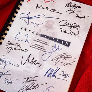 Puede incluir: Un guion encuadernado en espiral de la película "Interstellar" con numerosas firmas en varios colores. El título y los créditos son visibles, junto con una imagen fija de la película. El fondo es una tela roja texturizada.