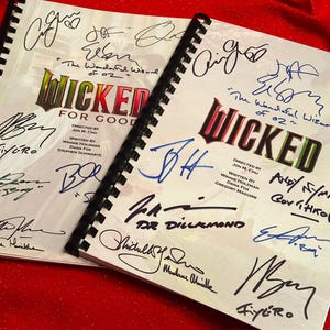 May include: Two spiral-bound scripts from the musical "Wicked" are displayed on a red surface. The covers feature the title in green and red, with numerous signatures in various colors. The text "For Good" is also visible.