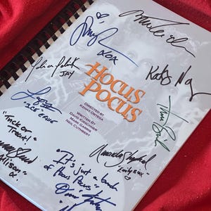 Op de afbeelding: Een wit boek met de titel "Hocus Pocus" en handtekeningen van de cast van de film. De handtekeningen zijn van Bette Midler, Sarah Jessica Parker, Kathy Najimy en anderen.