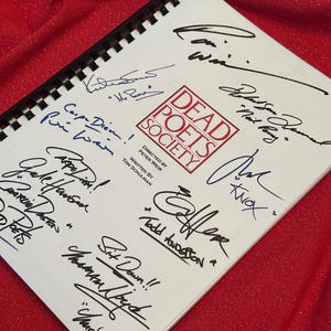 May include: A spiral-bound notebook with the title "Dead Poets Society" and numerous signatures in black ink. The cover features the film's logo in red. The notebook is on a red, textured surface.