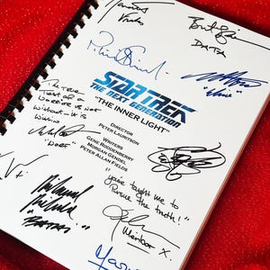 Puede incluir: Un cuaderno blanco con espiral con el logotipo de Star Trek: The Next Generation y el título "The Inner Light". El cuaderno está cubierto de firmas, con texto que incluye "The True Test of a Warrior is Not Without - it is Within."