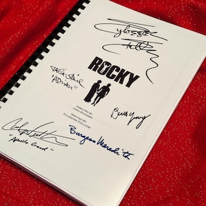 May include: A white script with the title "ROCKY" in black text. The script has signatures from the actors Talia Shire, Burt Young, and Carl Weathers. The signatures are in black ink. The script also includes the names of the director and writer, John Avildsen and Sylvester Stallone, respectively.