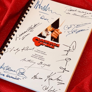 May include: A white notebook with a black spiral binding. The cover features a black and white illustration of a character from the film "A Clockwork Orange". The notebook is signed by the cast and crew of the film, including Malcolm McDowell, Stanley Kubrick, and Anthony Burgess.