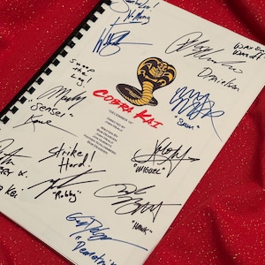 May include: A white page with a black and red Cobra Kai logo and the title "Cobra Kai" with the date "December 19". The page is signed by several people, including the actors who played Daniel LaRusso, Johnny Lawrence, and Miguel Diaz in the Netflix series.