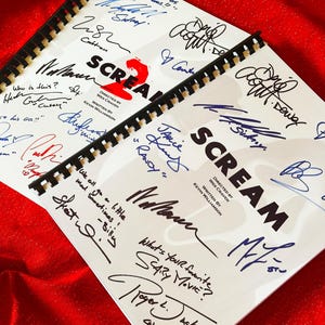 Könnte beinhalten: Zwei spiralgebundene Drehbücher der Filme "Scream" und "Scream 2" mit Unterschriften in verschiedenen Farben. Die Drehbücher befinden sich auf rotem Hintergrund. Das "Scream 2"-Drehbuch enthält den Text "Regie: Wes Craven, Drehbuch: Kevin Williamson".