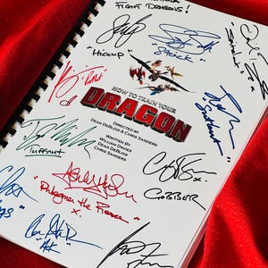 May include: A white book with a black spiral binding. The cover features a colorful illustration of dragons and the title "How to Train Your Dragon". The book is signed by several people, including the names "Hiccup", "Tuffnut", "Astrid", and "Gobber".