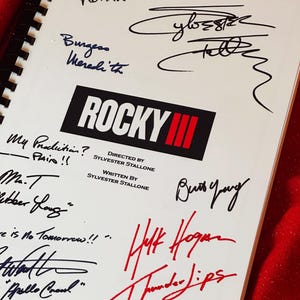 Könnte beinhalten: Ein weißer spiralgebundener Notizbuchdeckel mit dem Rocky III-Logo in Schwarz und Rot. Der Deckel zeigt mehrere Unterschriften und handschriftliche Zitate in schwarzer und roter Tinte, darunter Namen wie Sylvester Stallone, Mr. T und Hulk Hogan.