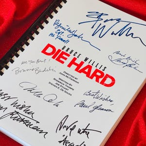 May include: A white book with the title "Die Hard" in red letters. The book is signed by Bruce Willis, John McTiernan, Paul Gleason, and other cast and crew members. The book is on a red surface.
