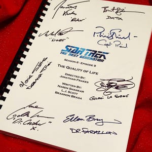 Puede incluir: Un guion firmado del episodio "The Quality of Life" de Star Trek: The Next Generation, temporada 6. El guion, encuadernado con espiral negra, presenta el logo de la serie y está firmado por el equipo, incluido Jonathan Frakes. Fondo rojo.