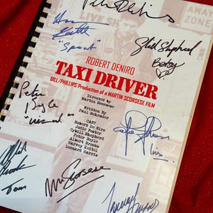 Puede incluir: Un guion encuadernado en espiral de la película "Taxi Driver" con múltiples firmas en tinta negra y azul. La portada presenta el título de la película en rojo y los nombres de Robert De Niro y Martin Scorsese.