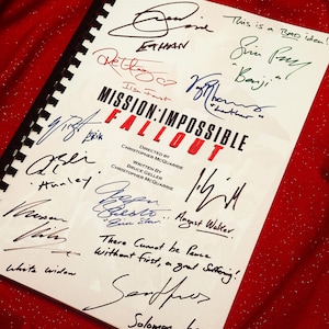 May include: A script for "MISSION: IMPOSSIBLE - FALLOUT" with multiple signatures in different colours. The title is in red and black. The script is spiral-bound and rests on a red, textured surface.