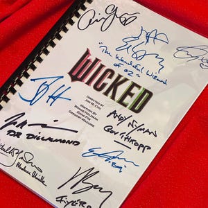 May include: A white book with the title "Wicked" in green and blue letters. The book is signed by several people, including "Andy Nyman" and "Gov't Thorpe".