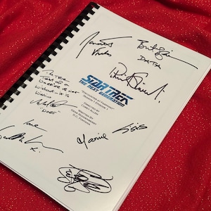 May include: A spiral-bound script for "Star Trek: The Next Generation" with multiple signatures in black ink. The title "Encounter at Farpoint" is visible, along with the names of Gene Roddenberry, Corey Allen, and D.C. Fontana. The script rests on a red, textured surface.