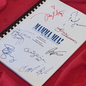 Può includere: Uno script bianco con il titolo "Mamma Mia! The Movie" e i nomi "Phyllidia Lloyd" e "Catherine Johnson" stampati su di esso. Lo script è firmato da diverse persone, tra cui "Julie Walters" e "Amanda Seyfried".