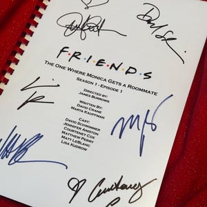 Pode incluir: Um roteiro da série de TV "Friends" com várias assinaturas em tinta azul e preta. O título "FRIENDS" está no centro, com detalhes do episódio abaixo. O roteiro está em papel branco com encadernação em espiral vermelha.