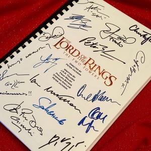 May include: A white page with a black spiral binding, featuring the title "The Lord of the Rings: The Two Towers" in gold lettering. The page is signed by multiple individuals, including Peter Jackson, the director of the film adaptation.