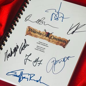 Puede incluir: Un cuaderno blanco con una encuadernación en espiral negra. La portada presenta el logotipo de la película Piratas del Caribe: La Maldición de la Perla Negra. El cuaderno está firmado por Johnny Depp, Orlando Bloom, Keira Knightley, Jack Davenport y Geoffrey Rush.