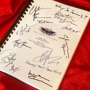 Może przedstawiać: Biały notatnik ze scenariuszem z czarną spiralną oprawą, zawierający liczne podpisy i tytuł "The Dark Knight". Na okładce widnieje logo Batmana oraz nazwiska reżysera i scenarzystów. Widoczne są również frazy "Why So Serious?" i "Joker".