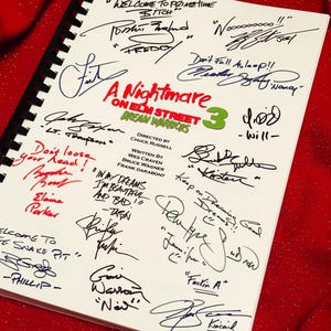 Op de afbeelding: Een spiraalgebonden script voor "A Nightmare on Elm Street 3: Dream Warriors" met talrijke handtekeningen in zwarte en blauwe inkt. De titel is in rood en groen, met het nummer "3" in groen. De achtergrond is een rode stof.