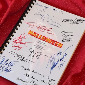 Peut inclure: Un livre blanc avec le titre "Halloween" en lettres rouges et noires. Le livre est signé par plusieurs personnes, dont Jamie Lee Curtis, qui a signé "Laurie" et "Sleep tight kids!!".
